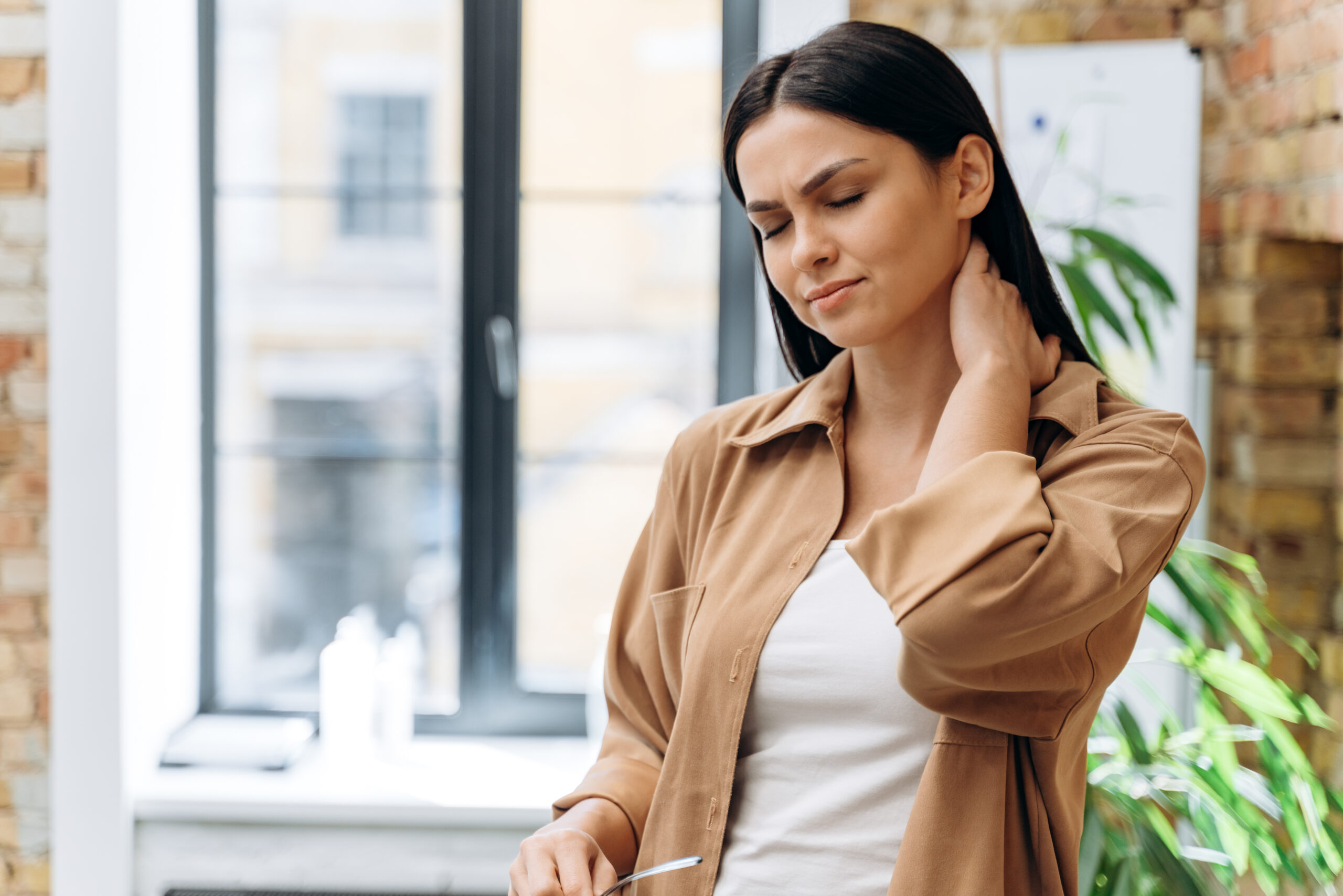Young businesswoman touching and massaging stiff neck to relieve pain in muscles after sedentary computer work in incorrect posture. Woman suffering from fibromyalgia at work